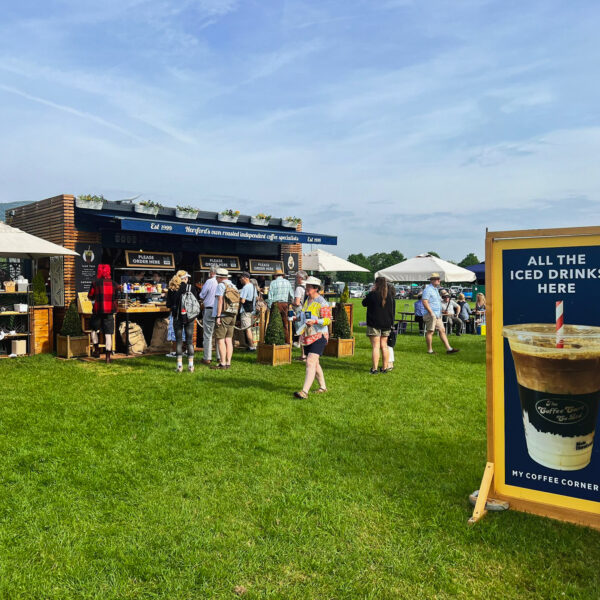 A bustling scene at an outdoor event kiosk at RHS Malvern 2024, with a crowd of people eagerly waiting for coffee. The kiosk is prominently displayed with a promotional board at the front, showcasing offers or information. The sky above is clear and blue, adding to the lively and vibrant atmosphere of the event.