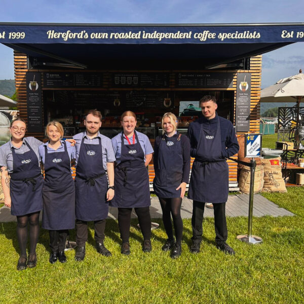 Six team members stand together in front of the event kiosk at the Malvern Spring Flower Show, RHS Malvern 2024. They are smiling and posed for the photo, with the kiosk serving as the backdrop. The outdoor event atmosphere highlights their presence at the show.