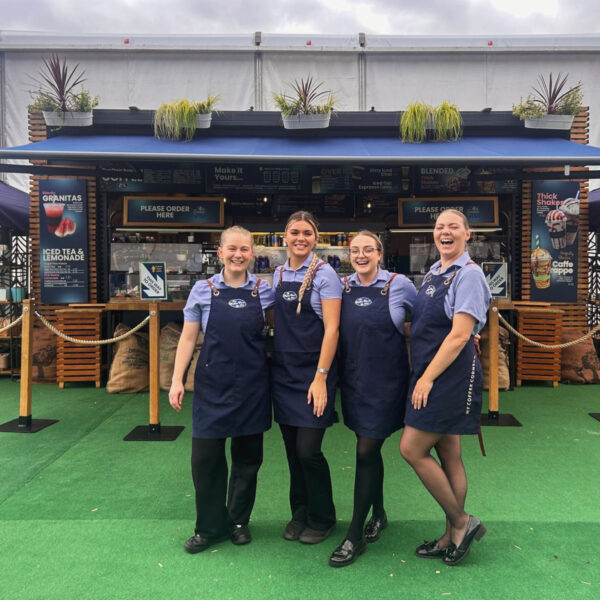 Four female staff members stand in front of the kiosk at the Longines Global Championships 2024 event, all smiling and happy. They are wearing their coffee cart uniforms and aprons, creating a friendly and professional appearance at the event.