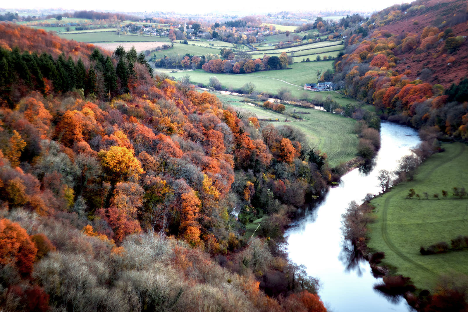 An aerial view of a lush valley during autumn, featuring a mix of colorful trees in shades of orange, red, and yellow. A serene river winds through the landscape, bordered by green fields and patches of forest. The scene captures a rural area with rolling hills in the background, dotted with small houses and farmland, creating a peaceful and vibrant autumnal atmosphere.