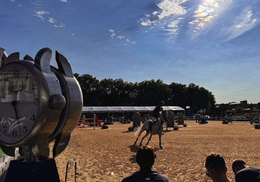 A horse and rider showcase their skills at the Longines Global Championships 2024 event, captured from the perspective of a nearby coffee kiosk. The rider navigates a course, demonstrating precision and agility, while spectators enjoy the event atmosphere in the background, creating a vibrant scene of equestrian excellence.