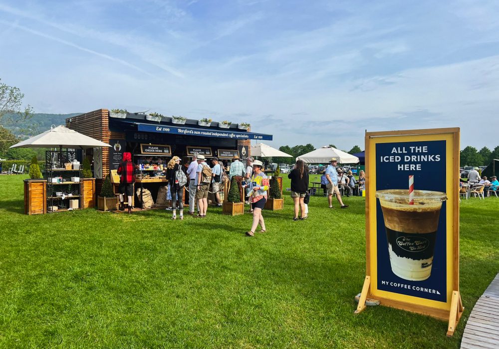 A bustling scene at an outdoor event kiosk at RHS Malvern 2024, with a crowd of people eagerly waiting for coffee. The kiosk is prominently displayed with a promotional board at the front, showcasing offers or information. The sky above is clear and blue, adding to the lively and vibrant atmosphere of the event.