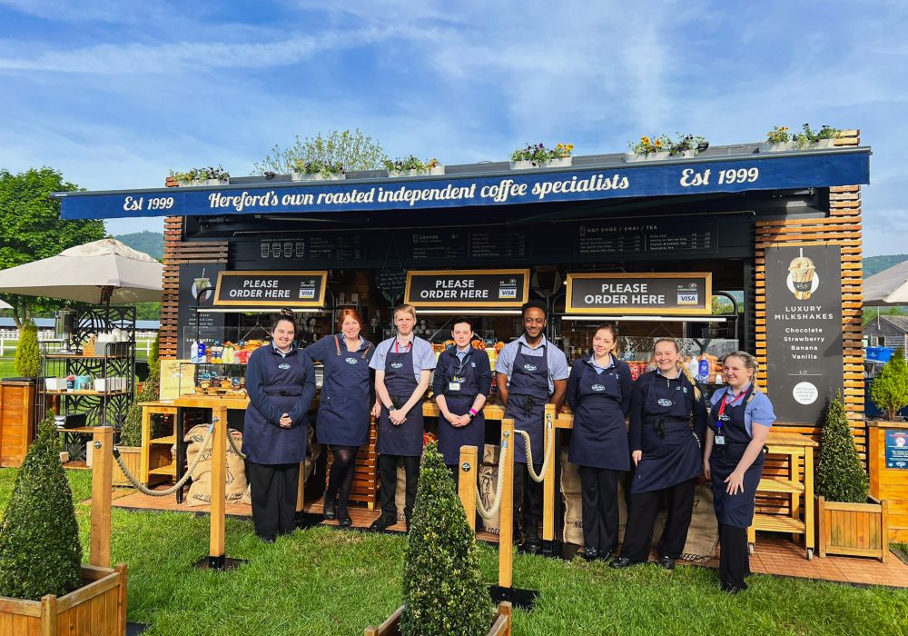 Eight members of the team stand in front of the event kiosk at the Malvern Spring Flower Show, RHS Malvern 2024. The team is posing together, smiling, with the kiosk as a backdrop. The setting is outdoors, with a lively event atmosphere, showcasing the team’s presence at the show.