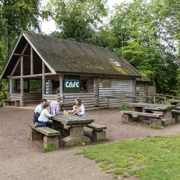 A small family sits at a wooden picnic table outside a rustic log cabin café, named 'Symonds Yat Rock Café.' The café is surrounded by tall green trees and a dirt path. Several more picnic tables are placed around the outdoor seating area, though they are unoccupied. The setting is peaceful and natural, with a relaxed atmosphere, blending well with the woodland surroundings.