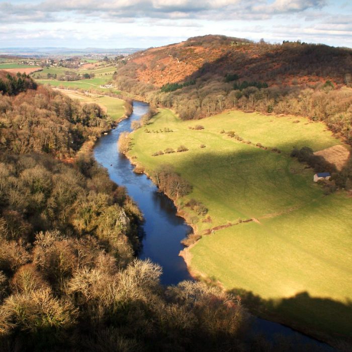 A sunny afternoon view overlooking the Wye Valley, with the landscape transitioning into autumn. Some of the trees have leaves turning orange, hinting at the season's change, while the valley remains lush and green under the clear sky, creating a serene and picturesque atmosphere.