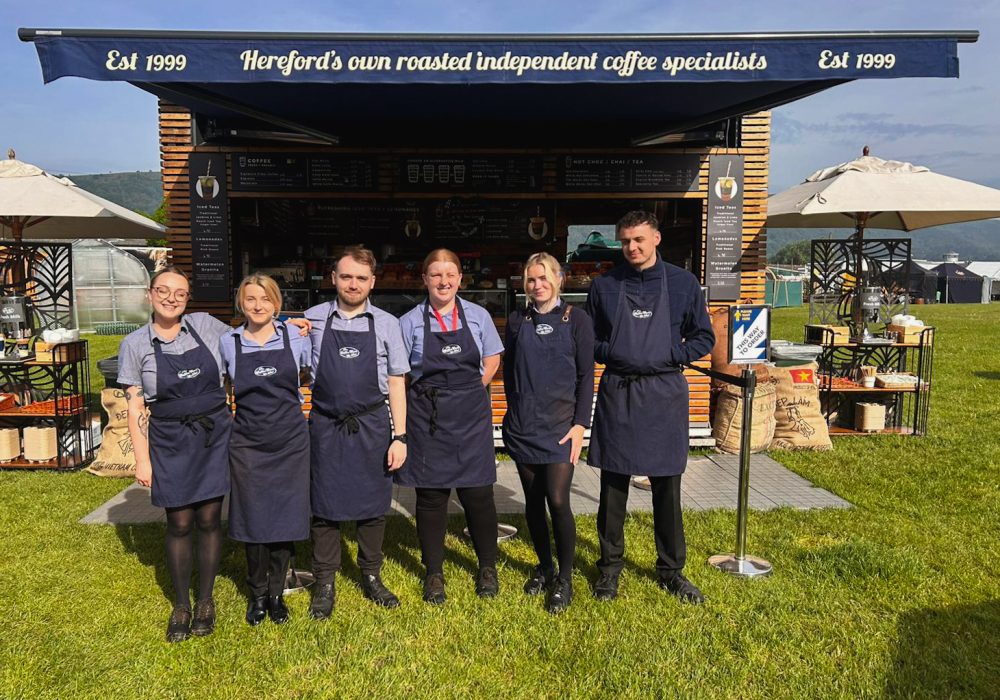 Six team members stand together in front of the event kiosk at the Malvern Spring Flower Show, RHS Malvern 2024. They are smiling and posed for the photo, with the kiosk serving as the backdrop. The outdoor event atmosphere highlights their presence at the show.