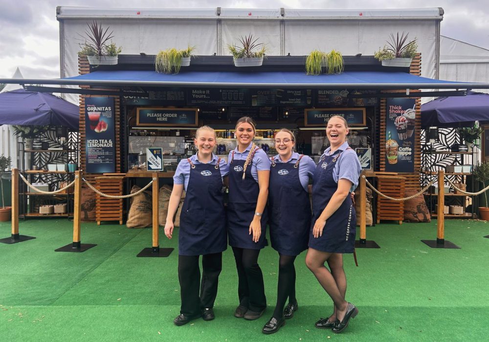 Four female staff members stand in front of the kiosk at the Longines Global Championships 2024 event, all smiling and happy. They are wearing their coffee cart uniforms and aprons, creating a friendly and professional appearance at the event.