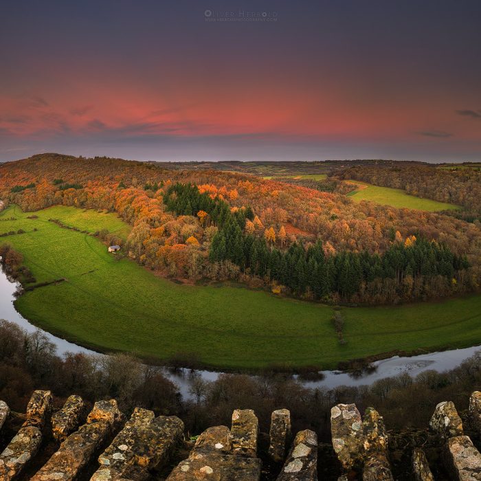 An aerial view of a lush valley during autumn at sunset, showcasing a vibrant mix of trees in shades of orange, red, and yellow. The golden light of the setting sun bathes the landscape, highlighting the rich autumn colors and casting long shadows across the valley, creating a warm and tranquil atmosphere.