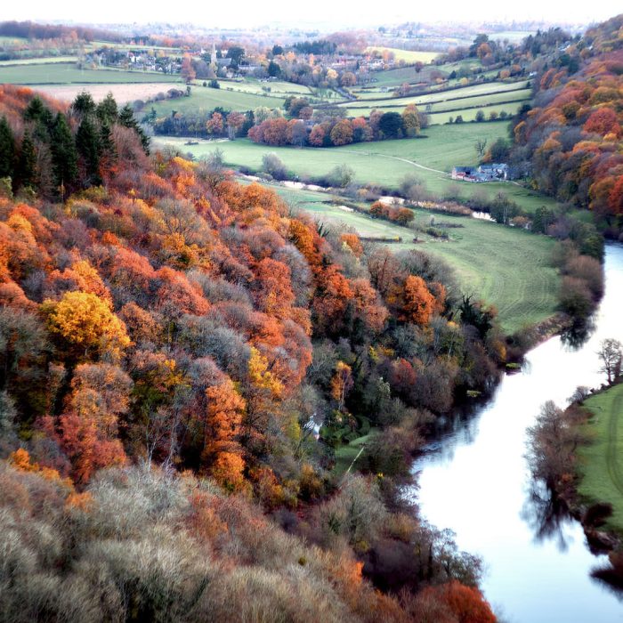 An aerial view of a lush valley during autumn, featuring a mix of colorful trees in shades of orange, red, and yellow. A serene river winds through the landscape, bordered by green fields and patches of forest. The scene captures a rural area with rolling hills in the background, dotted with small houses and farmland, creating a peaceful and vibrant autumnal atmosphere.