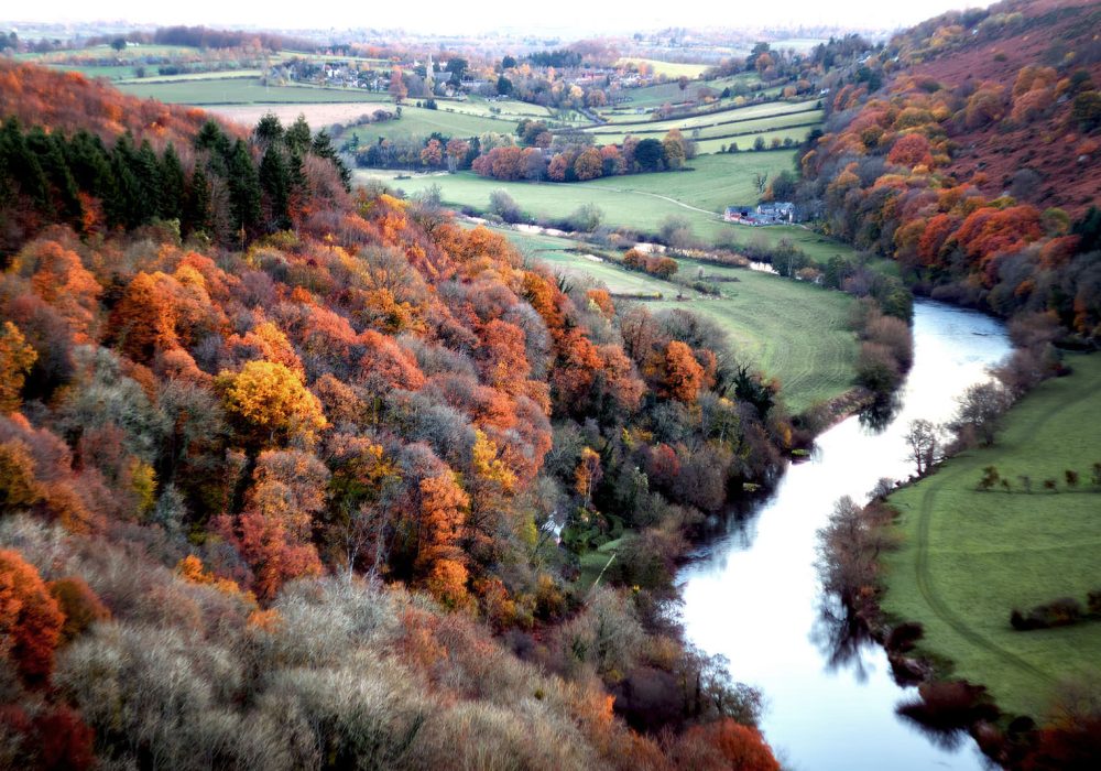 An aerial view of a lush valley during autumn, featuring a mix of colorful trees in shades of orange, red, and yellow. A serene river winds through the landscape, bordered by green fields and patches of forest. The scene captures a rural area with rolling hills in the background, dotted with small houses and farmland, creating a peaceful and vibrant autumnal atmosphere.